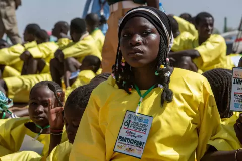 Sani Maikatanga A woman with beaded braids and a dark head covering looks at the camera. She is wearing a bright yellow top. Others in similar tops can be seen out of focus behind her.