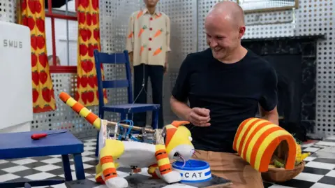 Nick Murdoch is kneeling and grinning as he watches the automaton cat dipping its head in a food bowl which has CAT printed on it. Nick is holding the orange and yellow striped tube which forms the machine's back. The cat has white paws and a white stomach. They are in the Plant Takeaway. There is a chair nearby with a screwdriver on it. There is a fridge with SMUG printed on it and a mannequin wearing a shirt printed with carrots. There is a mantelpiece and a pair of curtains with strawberries printed on them. On the wall is a print of the cat leaning over its food bowl.