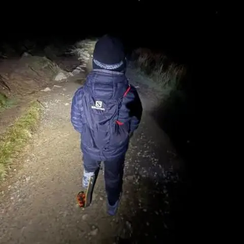 Daniel Thomas A picture of a young boy with a prosthetic limb on the left side on a mountain path in the dark. The image is taken from behind. He is wearing a navy padded coat and a black hat and the way in front of him is lit by a head torch that he is wearing. He is also carrying a navy rucksack. 