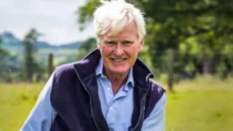 A man with white hair, wearing a blue jacket and light blue shirt