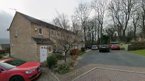 Google A quiet housing estate. A red car is parked to the left of the photo and more cars are visible in the distance. It is a grey day.
