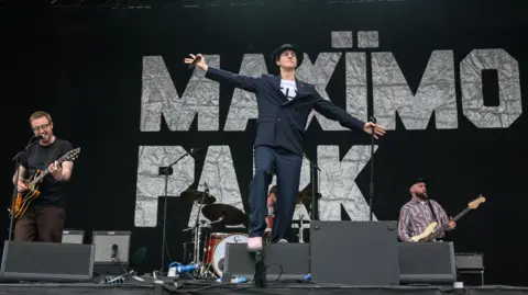 Katja Ogrin/Redferns Maximo Park on stage. Paul Smith is at the front with his arms outstretched. He is wearing a dark suit with the same hat as before. To the left of his is a man with dark, short hair playing an electric guitar and singing into a microphone. There is a drummer behind Paul Smith and another guitarist to the right of the drummer. There is a black sign which reads Maximo Park that covers the back of the stage.