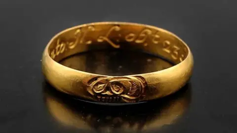 A gold ring resembling a wedding band reflecting on a polished black surface. It has a skull engraved on the top and an ornate inscription on the inside.