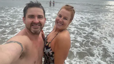 Anna Thomas A selfie of Dan (left) and his partner Anna (right) standing in the ocean. Dan has short, black hair, and Anna has red hair tied back. Both are smiling. There are also two people in the sea in the background.