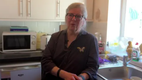 Josephine Wood, who has grey hair and black rimmed glasses and is wearing a blue shirt with a butterfly broch, speaks to the camera in the kitchen of a house.