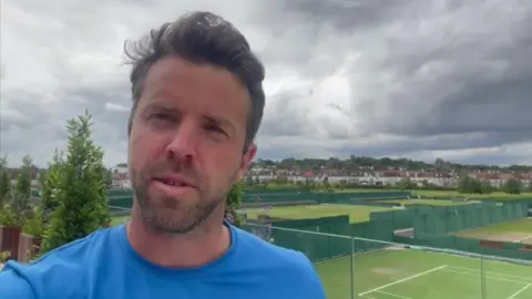 Richard Irwin, wearing a blue T-shirt, stands giving an interview with tennis courts in the background