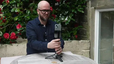 A man with a bald head and a beard, and glasses, holding his phone on a tripod on a table in his garden.