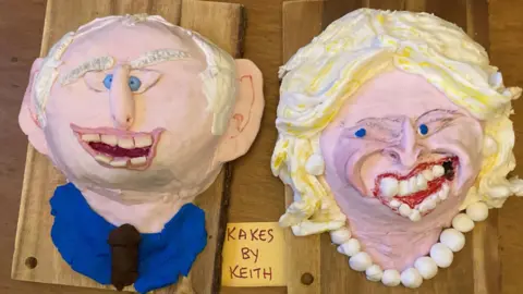 PA Media Two caricature cakes, one on the left showing a smiling King Charles, and one showing Queen Camilla with blonde hair and a very broad garish smile