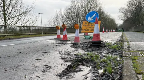 Kevin Shoesmith/BBC Grass and weeds grow in mud at the side of a road bridge. One lane is coned off. 