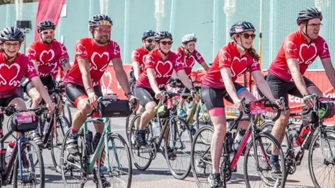 A crowd of cyclists all wearing red jerseys with a heart on the front cycle on a road