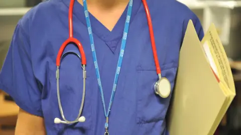 A medium shot of an unidentified doctor in uniform wearing an NHS lanyard and stethoscope around neck and carrying a file. 