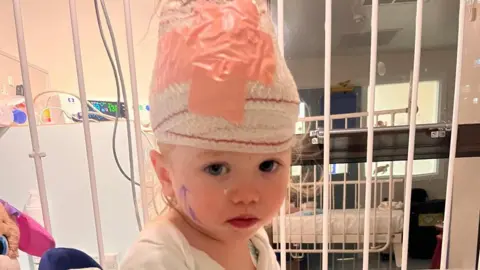 Family handout Tilly in hospital sitting up with bandages on her head.