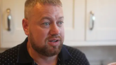 Steven Miller wearing a navy top with spots on it, pictured from the shoulders up. He has a tight beard and grey hair. There are cream coloured kitchen cabinets in the background. 