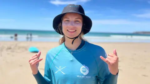 Vicky is standing on a sandy beach near the shoreline, wearing a blue long-sleeve rash vest and a dark wide-brimmed sun hat. She is making hand gestures with both hands. The ocean and a few distant beachgoers are visible in the background under a bright blue sky.