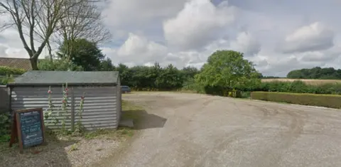 Google A gravel car park lined with hedgerow and trees with a shed in the foreground on a bright cloudy day
