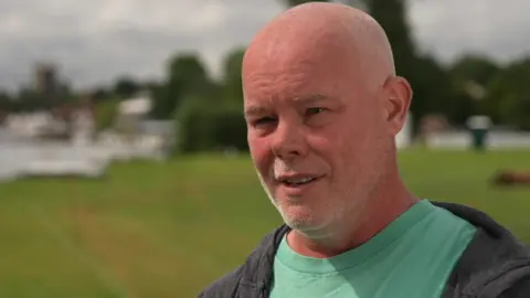 A bald Dave Wallace wearing a green T-shirt and dark hoody with a blurred field in the background