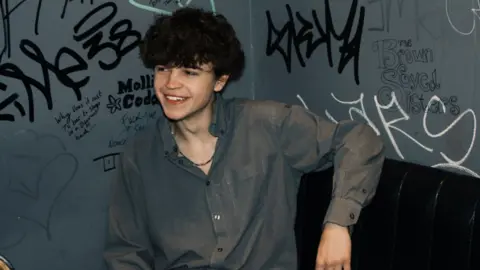 Cerys Longworth Tom A Smith, a young man with curly hair wearing a grey shirt unbuttoned to show a necklace, smiles into the distance while sitting on a black leather chair against a grey wall covered in graffiti.