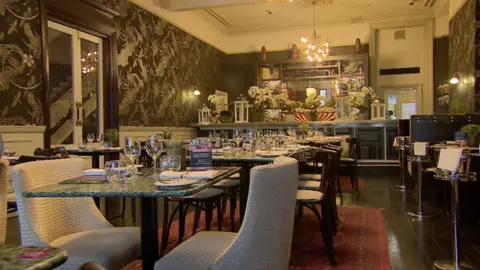 A dining room in a restaurant. The walls are adorned with dark, patterned wallpaper, while the ceiling is white. A light fitting hangs from the ceiling above the bar, at the far end of the room. In the foreground are green marble tables set with wine glasses and side plates. There are beige / light coloured fabric chairs pulled up to the tables. The dark wooden floor is covered by dark red rugs.