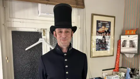 Jamie Tyler Jamie in a dressing room wearing a tall hat and a policeman's jacket, with a poster on the wall in the dressing room. He is standing in front of a window.