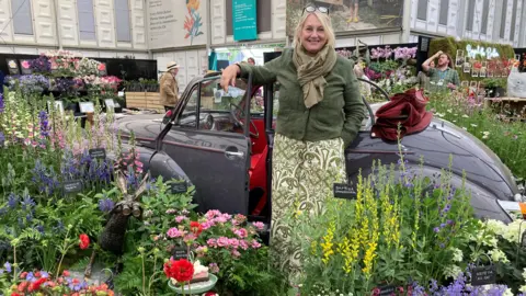 Stella Exley wearing a green jacket and a green and white long skirt stands in front of a Morris Minor with plants surrounding it at the Chelsea Flower Show.
