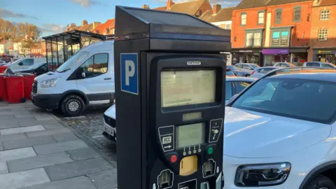 A parking ticket machine in the foreground with Yarm High Street in the background. Lots of vehicles are parked.