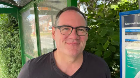 John Devine/BBC Andrew Copley has dark short hair, with grey flecks, he is wearing glasses with black frames and a dark V-necked T-shirt. Behind him is a bus stop shelter with timetable sign to the right.