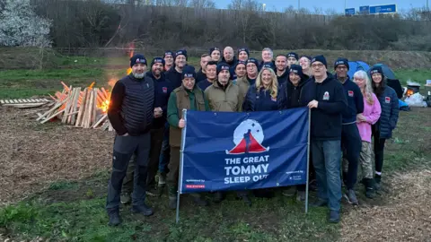 A group of men and women standing at a field, with a large blue banner which reads The Great Tommy Sleep Out. There is a campfire set up behind them.