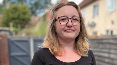 A woman with blonde hair and glasses looks at the camera, while standing in a back garden with fence panels behind her out of focus. She wears a black top.