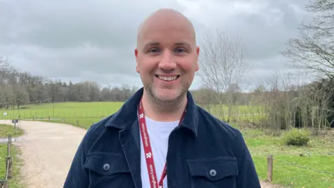 ELLEN KNIGHT/BBC Nick pictured looking into the camera and smiling. He's wearing a navy jacket over a white tshirt, and has a red lanyard with 'National Trust' written on it. He's stood on a gravel path, with green fields and trees seen in the distance. The sky is grey and cloudy. 