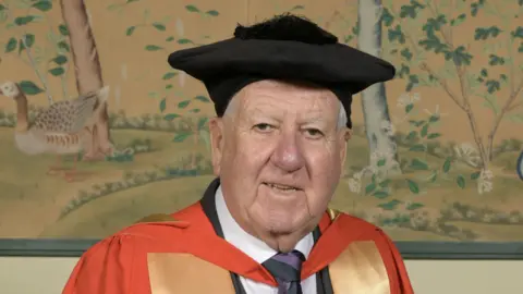Sir Bob Murray is wearing a black cap and red and yellow graduation clothes. He is wearing a purple and black striped tie as well as a white shirt. 