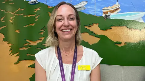 Clare Worden/BBC Emma Clark sits in front of a mural depicting the Norfolk coast in yellow, green and blue. Emma is wearing a white top with a purple lanyard and yellow name badge. She has blonde bobbed hair. 