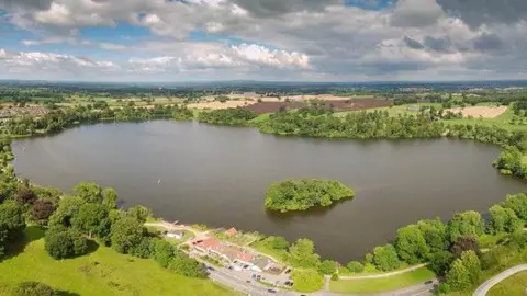 Shropshire Council A wide aerial picture of a large lake with a small tree-filled island in it. The lake is surrounded by trees, bushes and green fields as well as farmland.