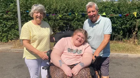 Sarah Davies (centre) with her parents on a tarmac surface in front of a hedge. Sarah is wearing a pink top and leopard print trousers, and is sitting in her powered wheelchair. Her mum Pam is standing to the left and wearing a yellow shirt and pale trousers, and her dad is standing to the right wearing a blue shirt and dark shorts.