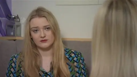 A BBC journalist with blonde hair wearing a blue and green patterned dress, looks at an interviewee who has the back to the camera. The interviewee is anonymous, so only the back of their head is visible. They also have blonde hair.