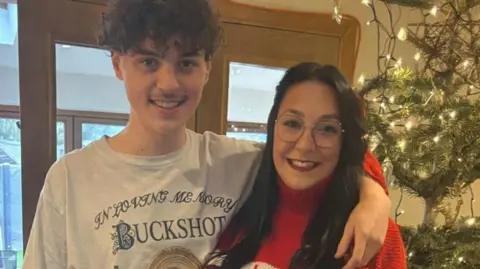 Corrin Wallace and her son Joshua are standing inside a house in front of windowed double-doors. There is a Christmas tree and lights in the corner of the photo. Corrin is wearing a red jumper and glasses. Joshua is wearing a white T-shirt with the words "Buckshot" on it. They are standing next to each other and he has his left arm over her left shoulder.