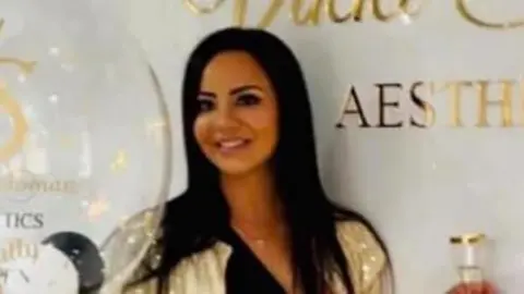 A person stands in a bright, modern interior beside a white wall featuring a gold logo and the text “AESTHETICS.” The person is wearing a black outfit with a gold belt and a gold sequined jacket, holding a large transparent balloon