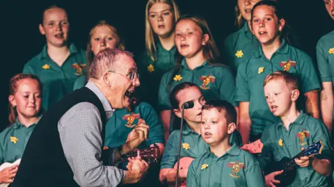 Aled Llywelyn | Eisteddfod Genedlaethol A male conductor sings towards a group of young children in matching green school uniforms as they sing.