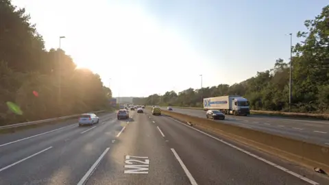 Google A Google street view screenshot of a four-lane motorway with cars and a lorry driving on it.