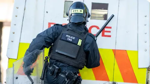 Aodhán Roberts Shows a police officer from behind. He is in full riot gear with a black helmet, black uniform and flak jacket. He is carrying a baton in his right hand and a shield in his left. He is standing in front of a white Land Rover, with yellow and orange stripes on it and the word police partially eclipsed by the officer's body. 