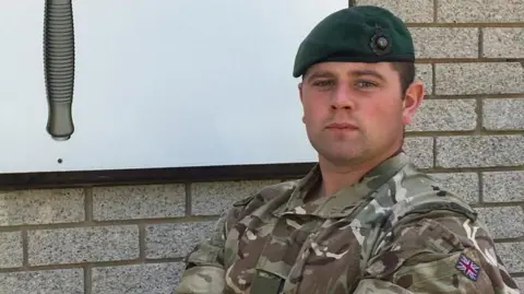 Danny Bowcock, who has his arms folded and is standing in front of a brick wall, is wearing a camouflage uniform and a dark green beret.