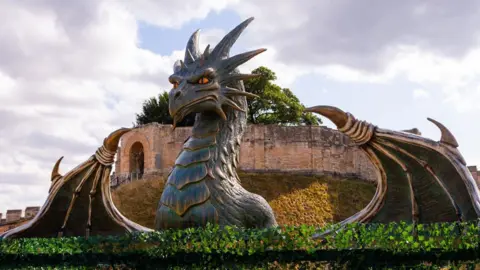 A large sculpture of a grey dragon with its wings outstretched is surveying the scene with striking yellow eyes. Castle walls are behind it.