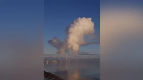 Bob Carter The picture shows a stretch of water, with an industrial site in the distance. Coming out of some of the towers are plumes of smoke. 