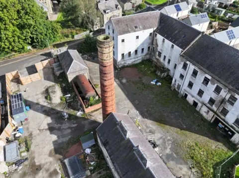 SOSE An old mill in the Borders views from above with a chimney rising into the sky and a white mill building in the background