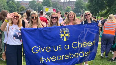 Five women are standing by a flag and most of them have one hand in the air. They are holding a large banner including the words "Give us a Cheer!" and a crowd of people and a building are behind them.