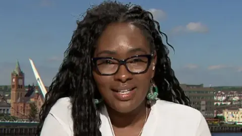 Lilian Seenoi Barr has long black hair and is wearing black-rimmed glasses.
She is wearing a white shirt and earrings.