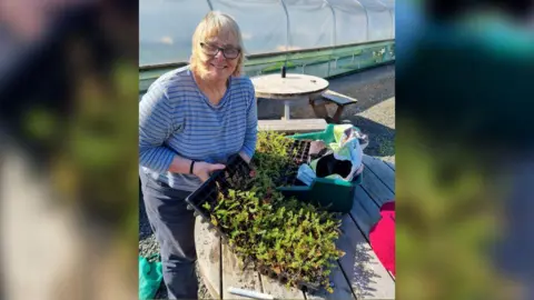 Woman with glasses and grey hair wearing a striped blue top and smiling by table with lots of plants on it. 