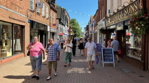 A pedestrianised street with a large number of people walking along it. There are shops either side, with hanging baskets and bunting also visible. Some of the shops have A-boards outside of them.