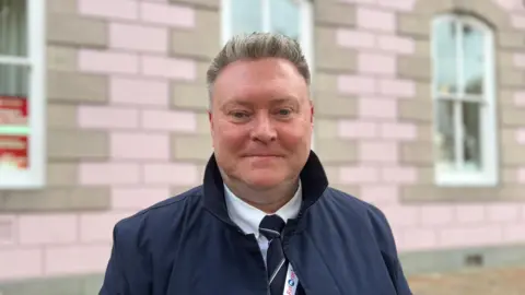 Deputy Rob Ward smiling at the camera. He is stood in front of a brick building. He is wearing a black coat and is wearing a white shirt with a striped tie. The building behind him is light brown and pink. 
