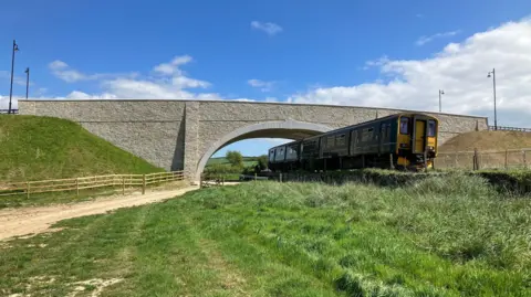 Network Rail A train going under the new bridge. The bridge is clad in granite. There is grass and new wooden fencing near the train track. There are four lamp posts on the road leading across the bridge.