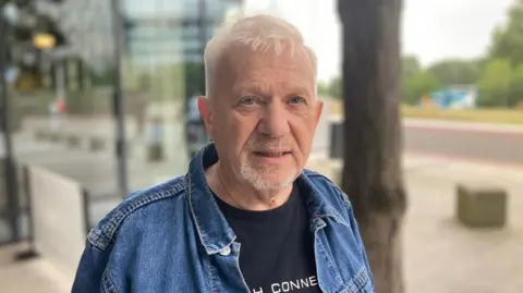 A man in a blue denim jacket and black top. Trees and a building are blurred in the background.